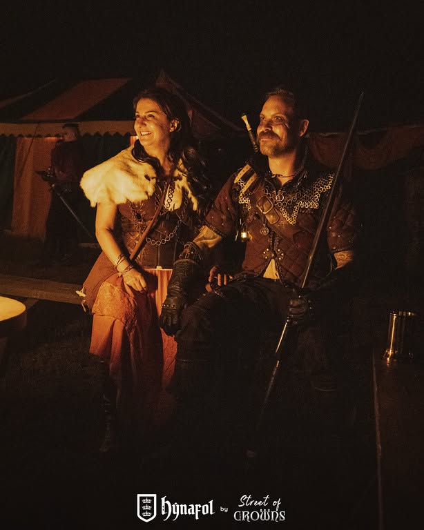 Talia and Korsin together in costume at Hynafol, lit by warm light in front of a medieval tent.